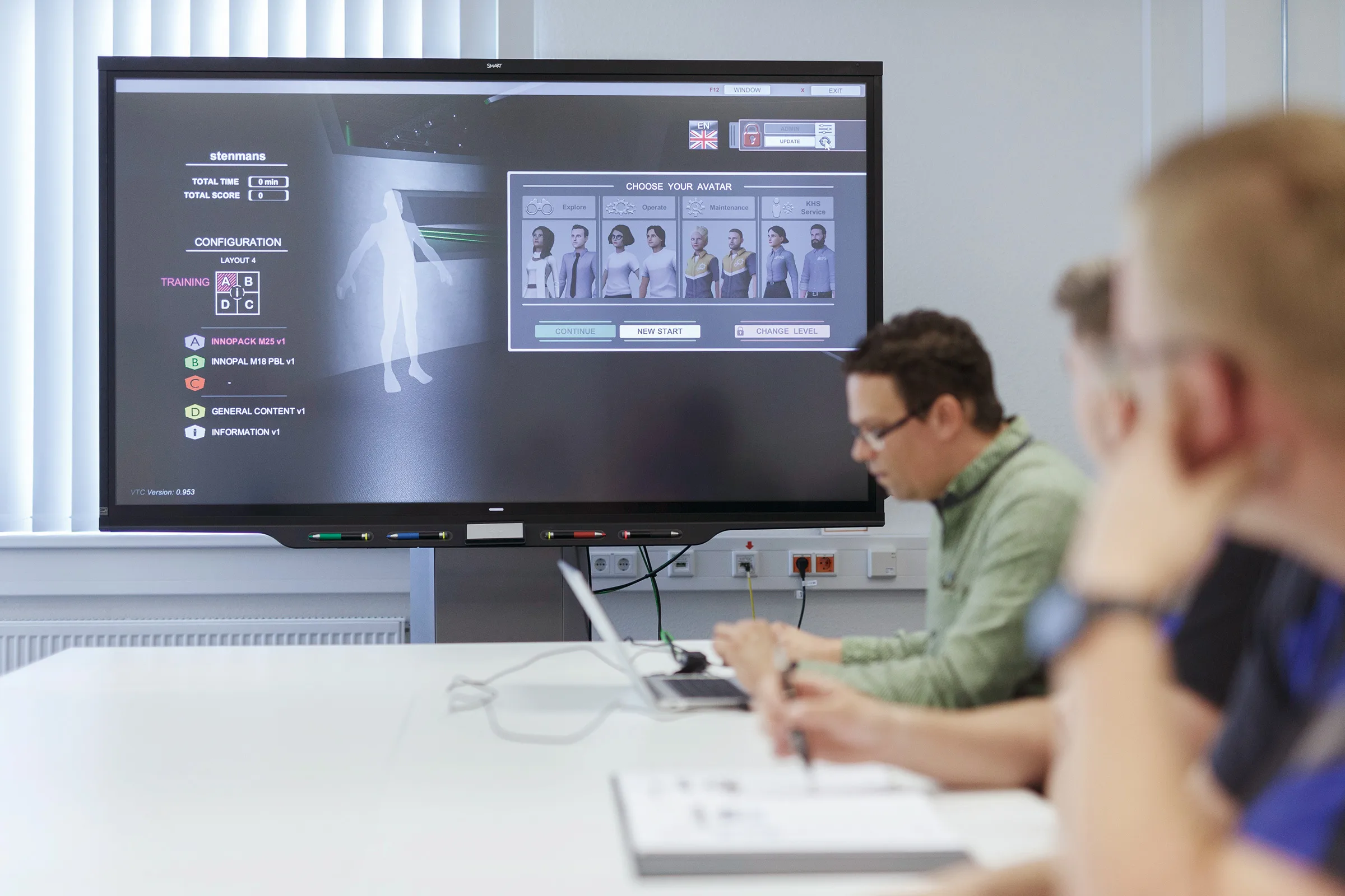 Un grupo de personas sentadas en una mesa con computadoras portátiles y una pantalla grande.